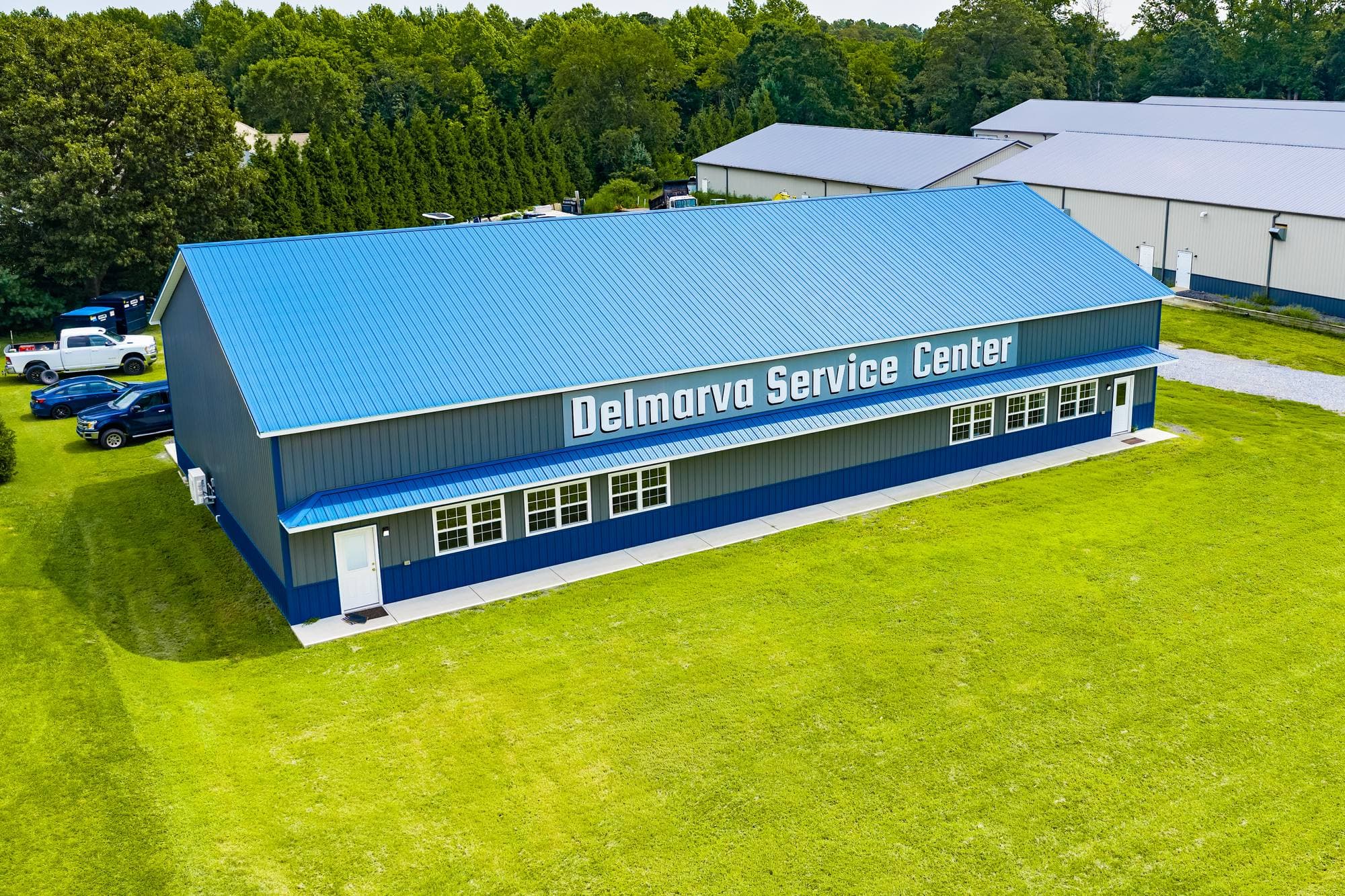 Aerial view of the Delmarva Service Center building and yard from the parking side, with work trucks parked beside the blue-and-grey shop